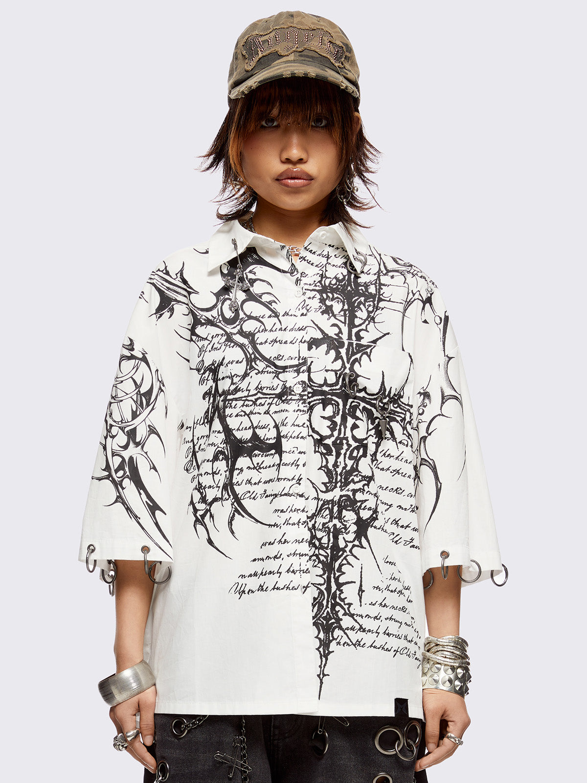 White oversized bowling shirt with black sigilism and lettering graphic print, eyelets with silver O-rings on sleeves and collar and more metal hardware details.
