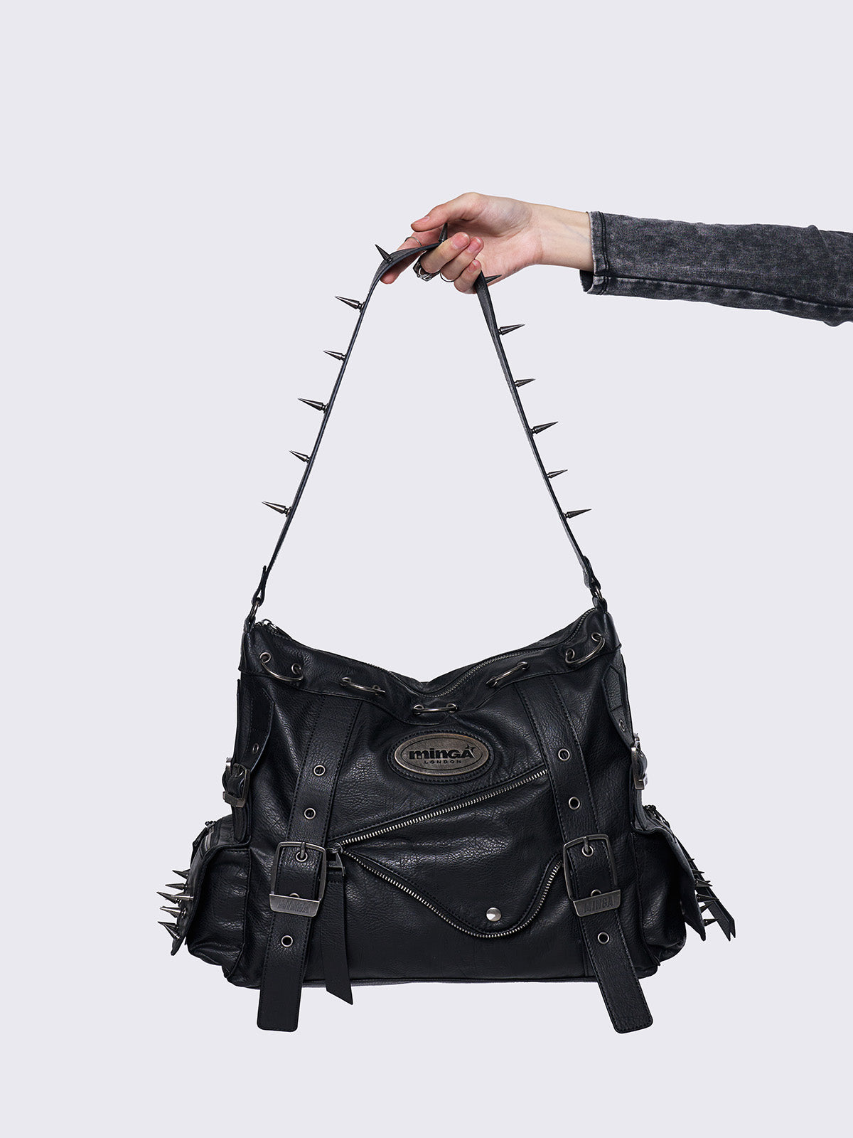 Black leather handbag with silver spikes on the strap, held by a hand, featuring buckle details and a rugged, edgy design.