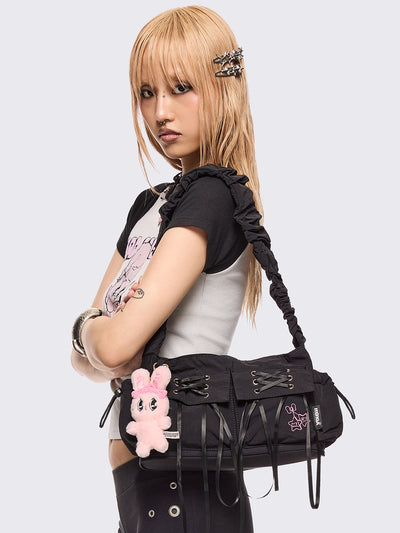 Black shoulder bag with ruched strap, lace-up ribbon front pockets, detachable plush pink bunny charm, and multiple pockets.