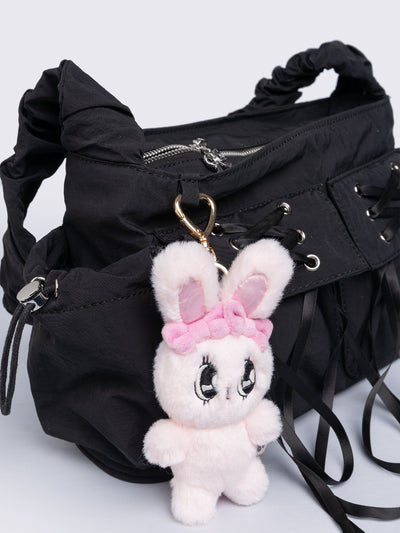 Black shoulder bag with ruched strap, lace-up ribbon front pockets, detachable plush pink bunny charm, and multiple pockets.