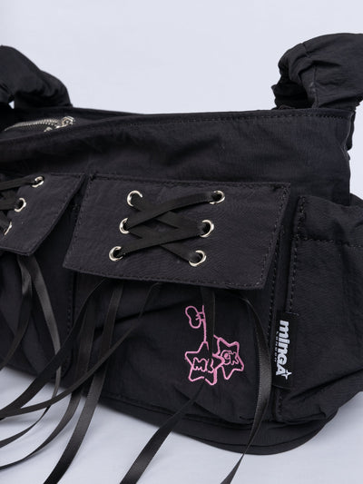 Black shoulder bag with ruched strap, lace-up ribbon front pockets, detachable plush pink bunny charm, and multiple pockets.