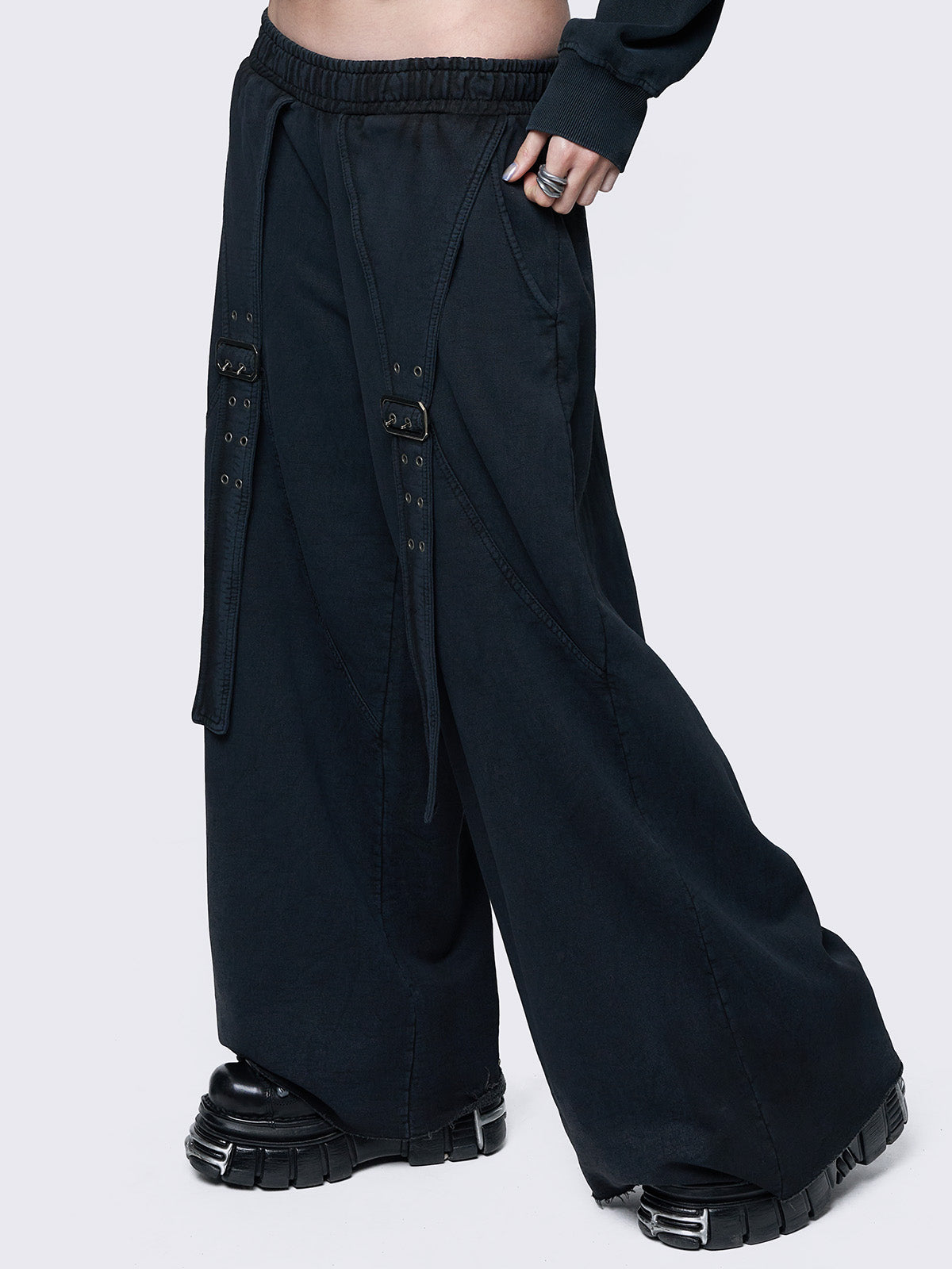 Wide-leg black joggers featuring adjustable buckle straps with grommet details, an elastic waistband, and an oversized fit. Designed for comfort and streetwear style.