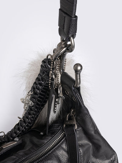 A black leather hobo bag decorated with silver studs, metallic charms, braided leather, and a large grey and white fur tail charm.