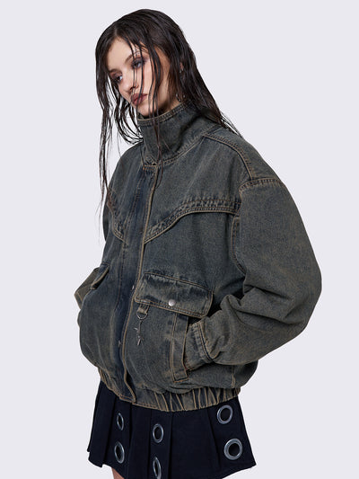 Blue washed denim bomber jacket with a brown overdye finish. The jacket features a boxy, oversized fit and a high crossover collar neck. It has multiple pockets and a Minga logo metal detail for added style.