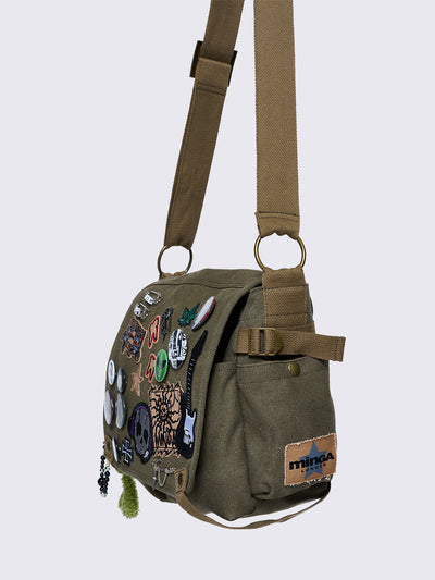 Olive green canvas messenger bag with grunge-inspired patches and pins, featuring a guitar, skull, alien, and other edgy designs. Includes a frog plush keychain and adjustable strap for a Y2K, alternative look.