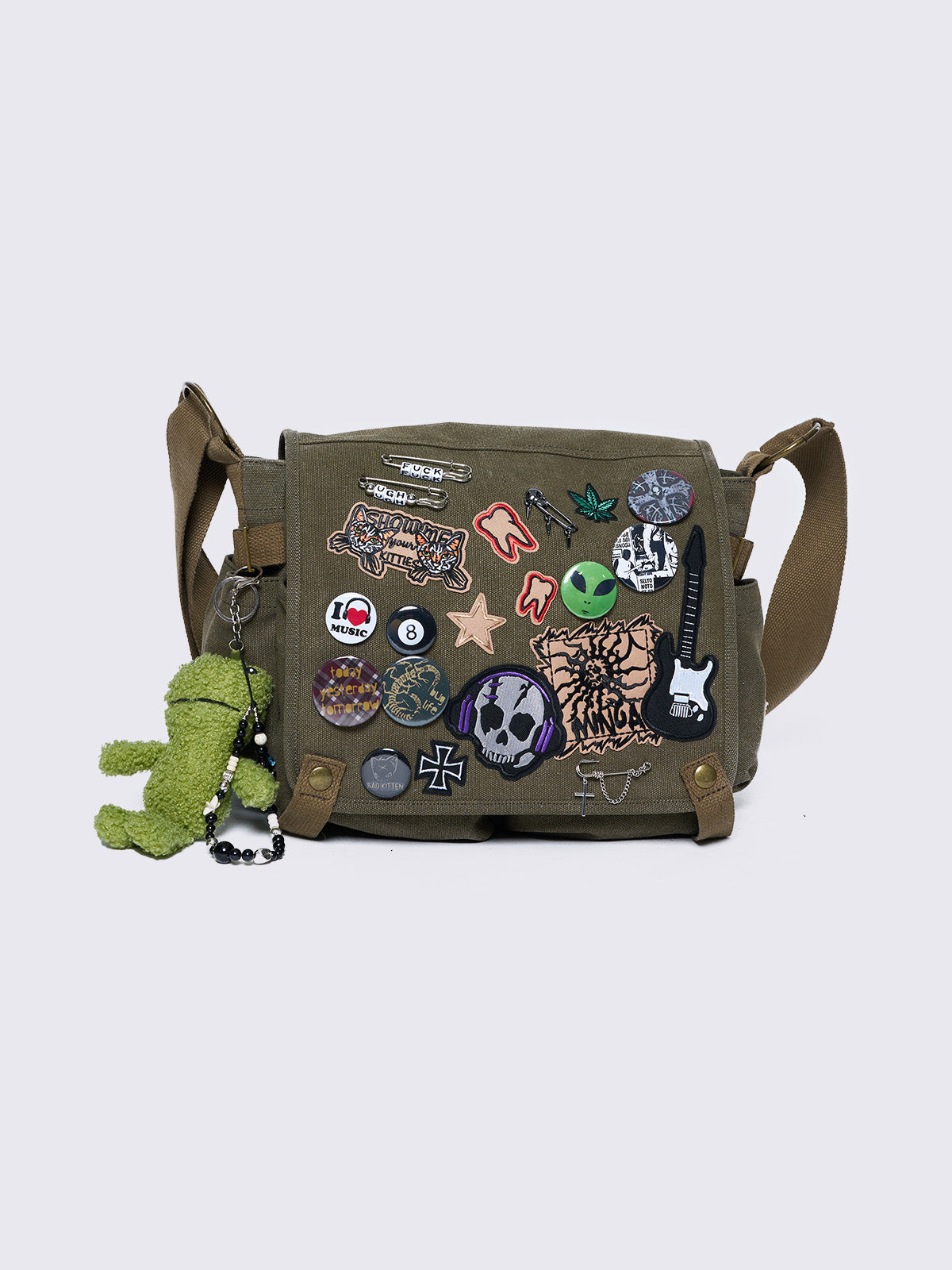 Olive green canvas messenger bag with grunge-inspired patches and pins, featuring a guitar, skull, alien, and other edgy designs. Includes a frog plush keychain and adjustable strap for a Y2K, alternative look.