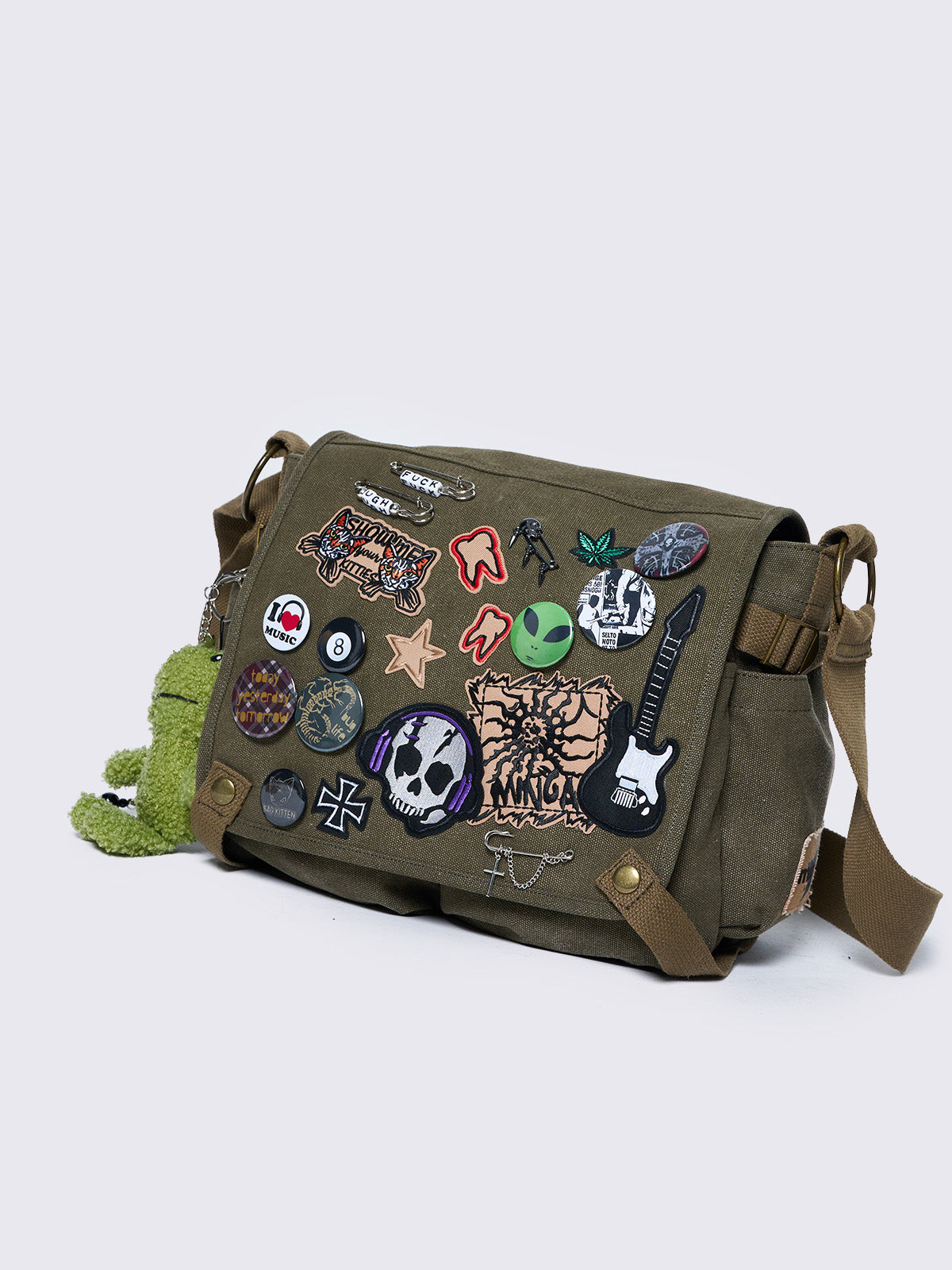 Olive green canvas messenger bag with grunge-inspired patches and pins, featuring a guitar, skull, alien, and other edgy designs. Includes a frog plush keychain and adjustable strap for a Y2K, alternative look.