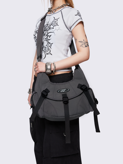 Grey nylon crossbody bag featuring multiple pockets with black webbing strap details. Flap and zip closures. Edgy, utility-inspired streetwear style.