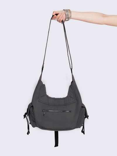 Grey nylon crossbody bag featuring multiple pockets with black webbing strap details. Flap and zip closures. Edgy, utility-inspired streetwear style.