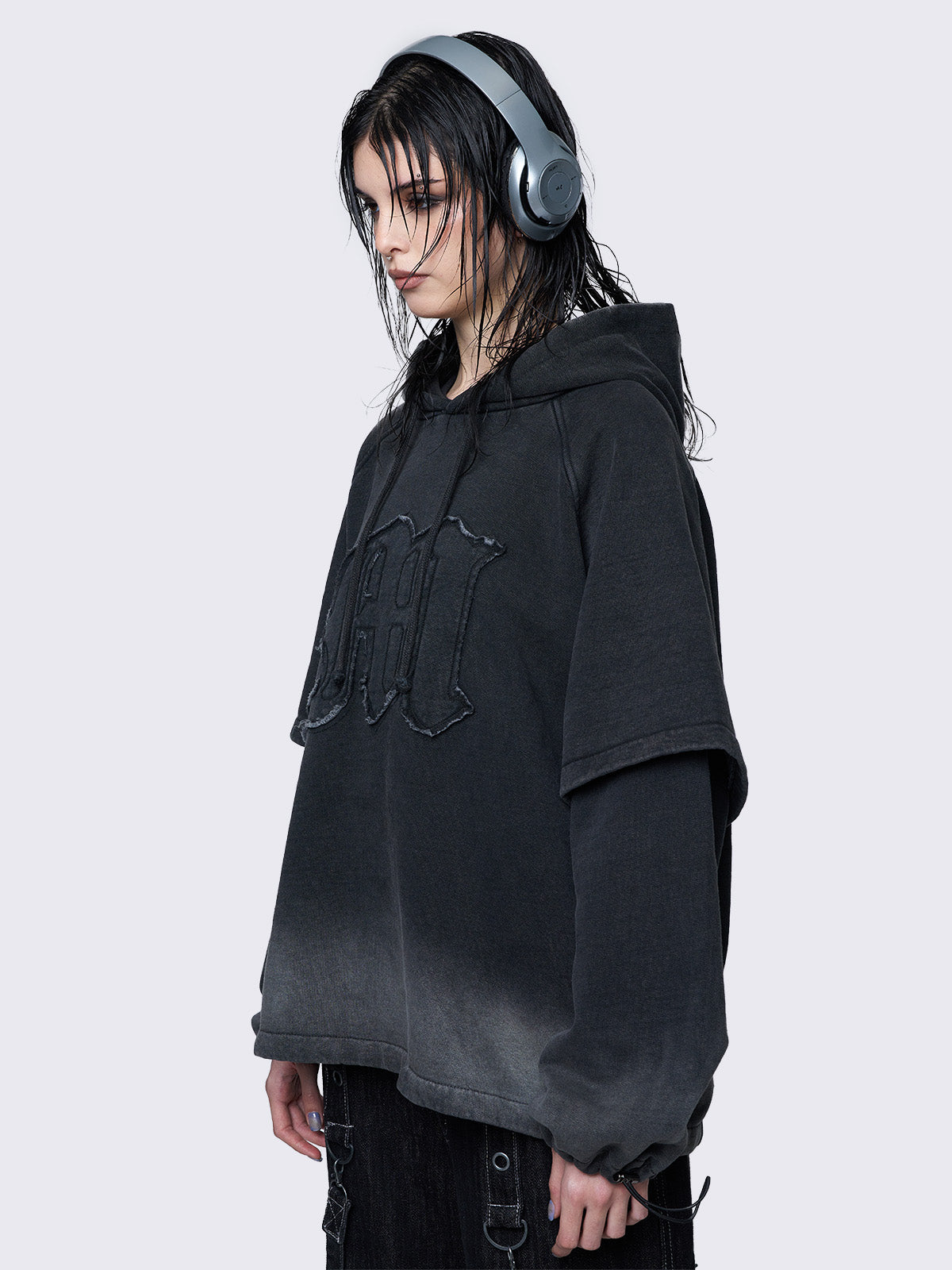 Oversized layered hoodie in washed black, featuring an embroidered "M" patch on the front, faded cuffs and hem, a drawstring hood, toggle elastic drawstrings, and raglan long sleeves.