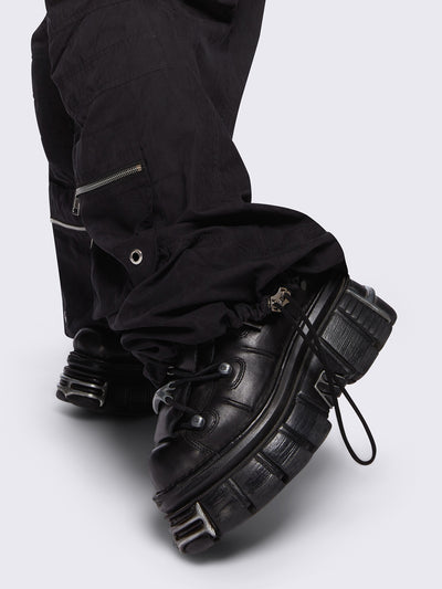 Black tech cargo parachute pants featuring multiple utility pockets with zipper and carabiner hardware detail - edgy, utility-inspired streetwear style.