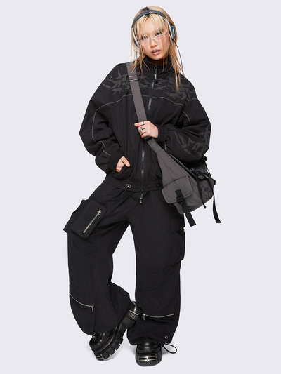 Black oversized windbreaker jacket with grey tribal graphics on the chest and sleeves, elastic cuffs, front zip closure, toggle-adjustable hem, and snap-button pockets  — edgy, utility-inspired streetwear style.