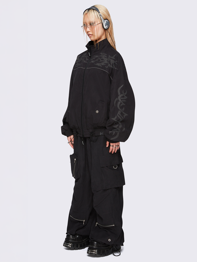 Black oversized windbreaker jacket with grey tribal graphics on the chest and sleeves, elastic cuffs, front zip closure, toggle-adjustable hem, and snap-button pockets  — edgy, utility-inspired streetwear style.