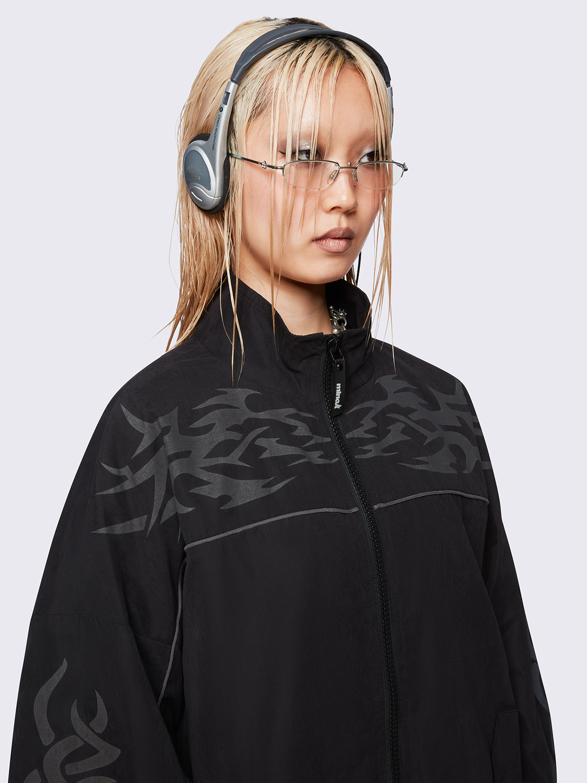 Black oversized windbreaker jacket with grey tribal graphics on the chest and sleeves, elastic cuffs, front zip closure, toggle-adjustable hem, and snap-button pockets  — edgy, utility-inspired streetwear style.