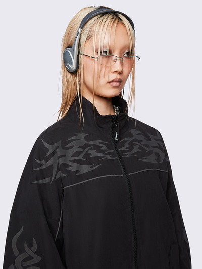 Black oversized windbreaker jacket with grey tribal graphics on the chest and sleeves, elastic cuffs, front zip closure, toggle-adjustable hem, and snap-button pockets  — edgy, utility-inspired streetwear style.