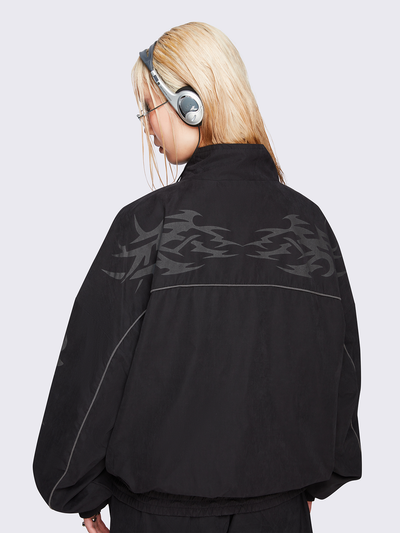 Black oversized windbreaker jacket with grey tribal graphics on the chest and sleeves, elastic cuffs, front zip closure, toggle-adjustable hem, and snap-button pockets  — edgy, utility-inspired streetwear style.