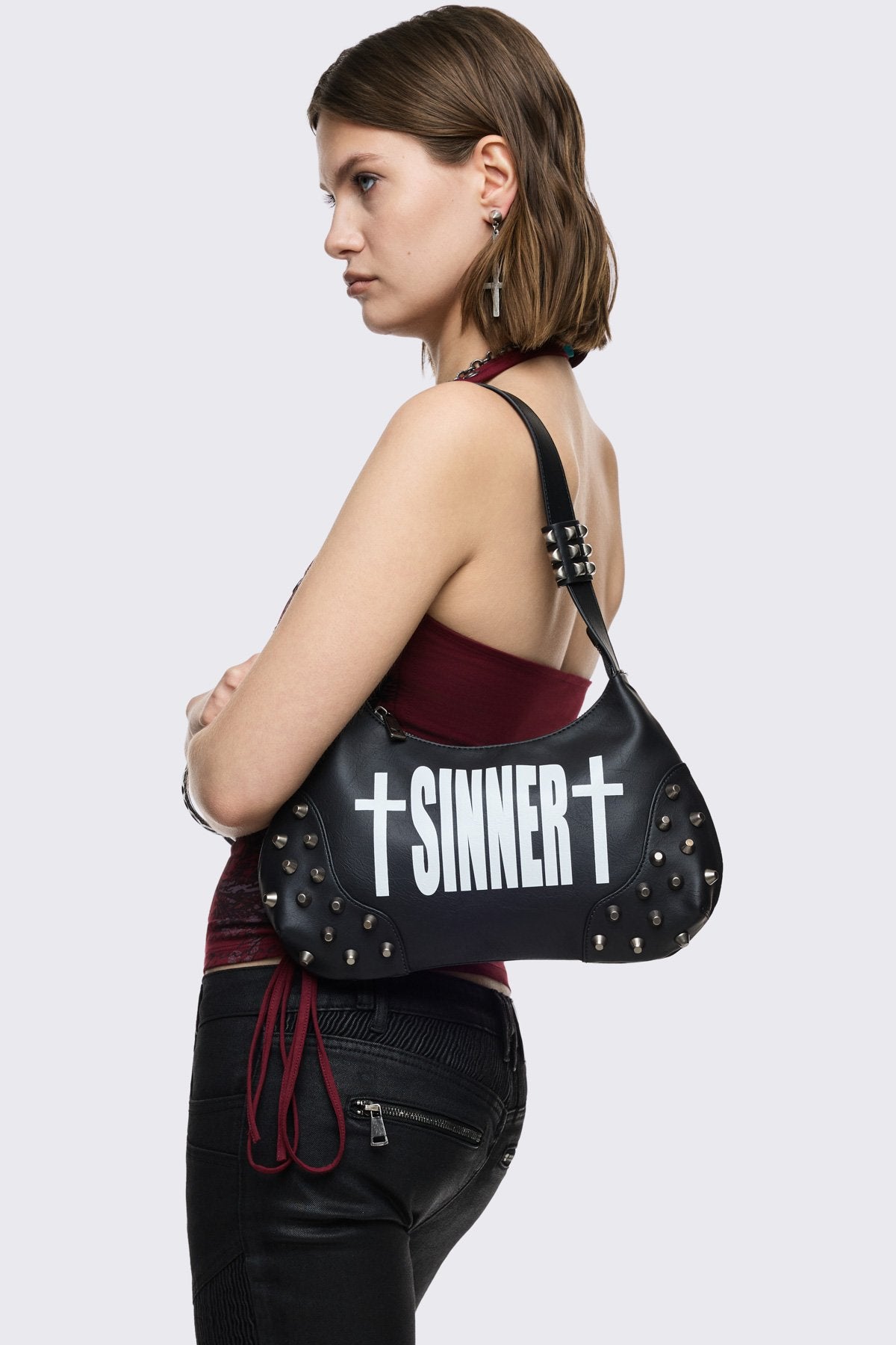Black faux leather shoulder bag with white sinner cross print, studded panels and grunge aesthetic.