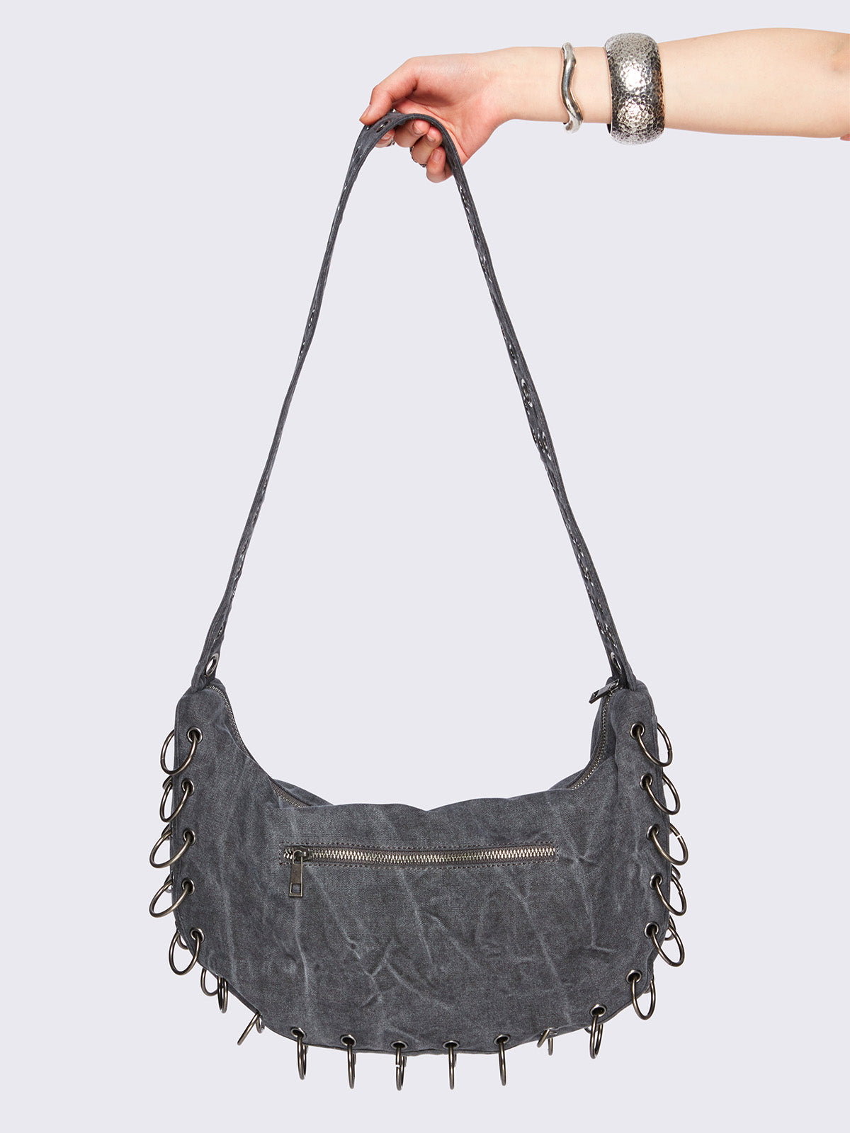 Grey washed canvas crossbody bag with a wide grommet strap, metallic logo patch, O-ring lace-up detailing along the edges, and zip-top closure.