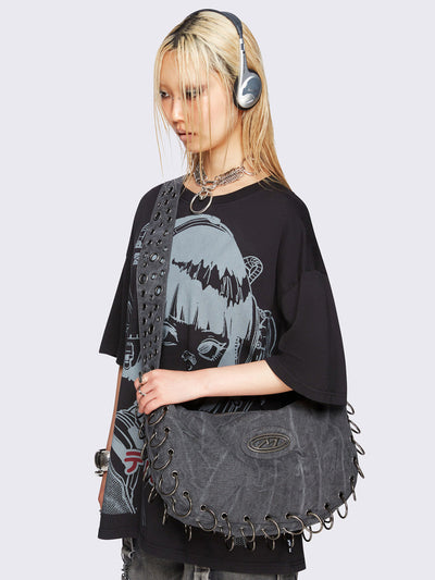 Grey washed canvas crossbody bag with a wide grommet strap, metallic logo patch, O-ring lace-up detailing along the edges, and zip-top closure.