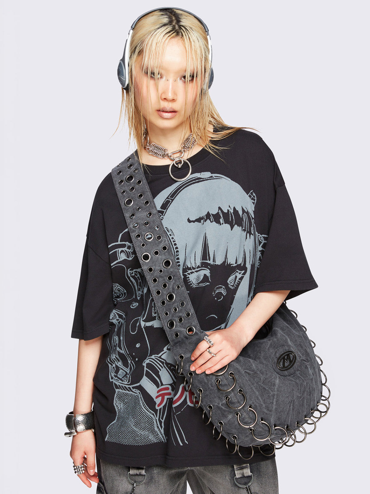 Grey washed canvas crossbody bag with a wide grommet strap, metallic logo patch, O-ring lace-up detailing along the edges, and zip-top closure.