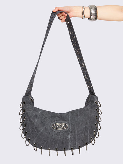 Grey washed canvas crossbody bag with a wide grommet strap, metallic logo patch, O-ring lace-up detailing along the edges, and zip-top closure.