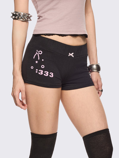 Black sweat micro shorts featuring kaomoji ASCII-style graphic print on side and back and a front pink bow detail. Low rise and elasticized waist.